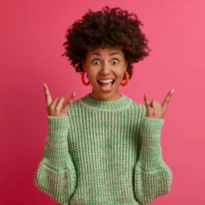 happy carefree dark skinned rebellious young woman enjoys awesome music makes rock n roll gesture has fun on music festival or cool event wears casual jumper poses against pink wall HyperMmedia